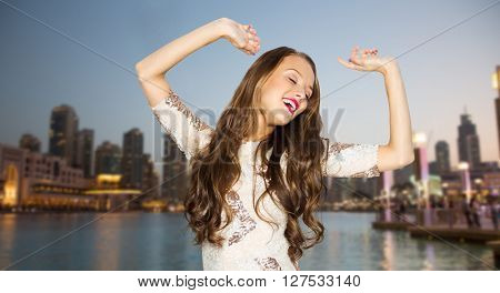 people, travel, tourism, holidays and fashion concept - happy young woman or teen girl in fancy dress with sequins and long wavy hair dancing at party over evening city waterfront background
