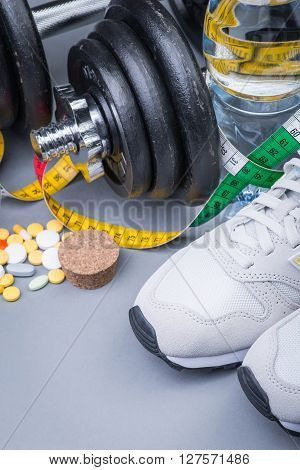 Dumbbells with measuring tape trainers pills and bottle of chilling water on grey