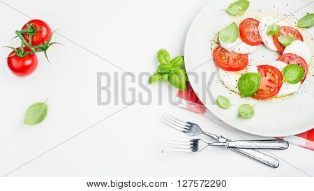 CLassic Italian Caprese salad with tomatoes, mozzarella di Buffala and fresh basil on red and white checkered napkin over white wooden background. Top view, copy space