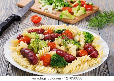 Delicious Spiral Pasta salad with broccoli and grilled sausages on a white dish with fresh tomato and sorrel green salad spring onions on a cutting board on the background close up