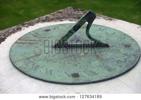 Sundial on plinth. Probably made from brass as it has tarnished green. Not enough sun to show the time. Backgound is lawn grass.