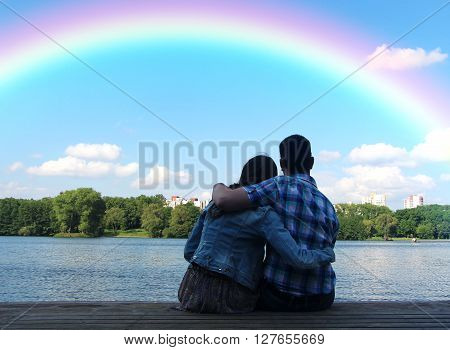 Two young man and woman looking at the rainbow on the background of the urban landscape and blue sky with clouds.