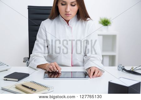 Doctor sitting at table with hands on tablet notebook and stethoscope aside. Front view. Concept of work.