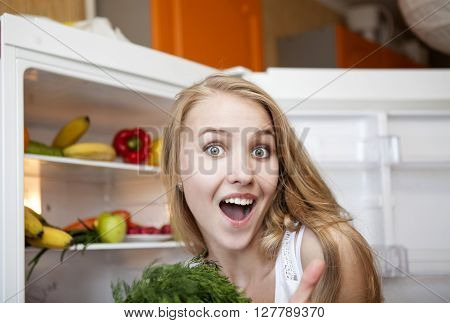 Beautiful surprised girl with open mouth opens the refrigerator and pulls fruits and vegetables