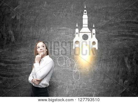 Startup concept with thinking businesswoman standing against chalkboard with rocket sketch