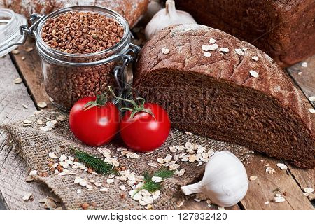 Close up photo of rustic bread on sackcloth, garlic, dill, two tomatoes, oat flakes and jar with buckwheat on planked wooden table