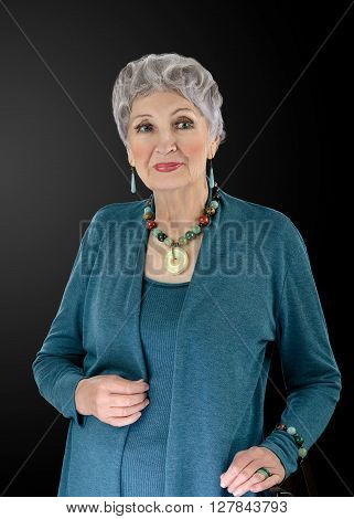 Portrait of spectacular 76-year-old woman wears multi colored semiprecious stone beads necklace with donut pendant and bracelet. Grey haired woman is clothed in old turquoise shrug set. Upper body vertical shot on black background
