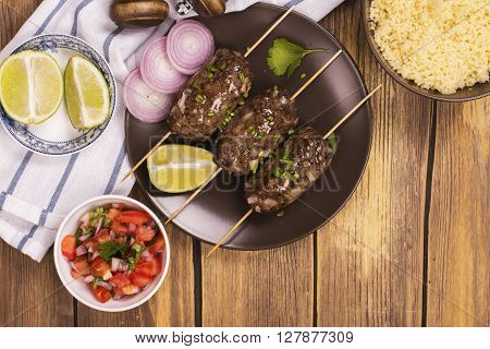 Grilled kebabs with vegetable salad, couscous and seasonings over wooden table