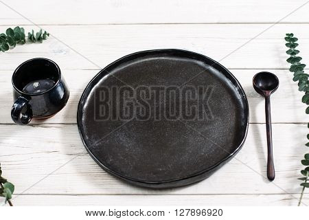 Top view empty black dishes and cutlery on rustic wooden table. Ceramic black plate, cup and spoon on white wooden background. Flat lay of handmade black ceramics on white wooden table.
