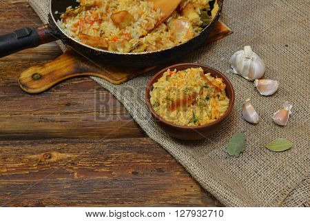 Vintage clay bowl with traditional spicy food pilaf, frying pan with pilaf on wooden background and the burlap. Pilaf with meat