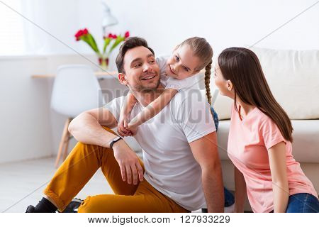 Involved in joy. Cheerful pretty little girl holding on the back of her father while spending time together with the family