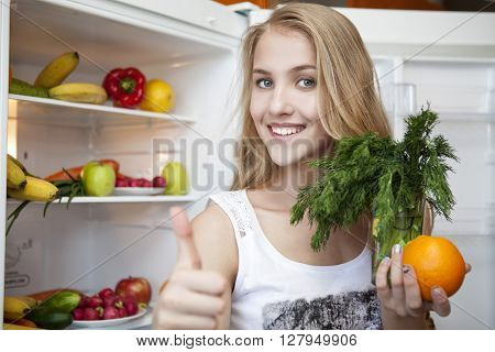 Beautiful young girl with thumb upward opens the refrigerator and pulls fruits and vegetables