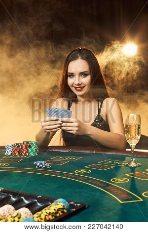 Gorgeous Emotional Young Woman In A Black Dress Sitting At Poker Table With Glass Of Champagne. Let 