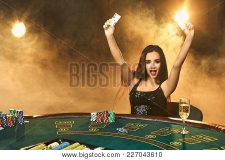 Gorgeous Emotional Young Woman In A Black Dress Sitting At Poker Table With Glass Of Champagne. Let 
