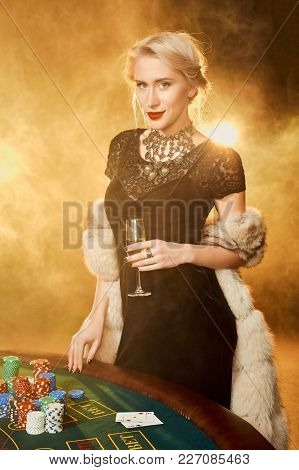 Young Woman In Evening Dress With Chips In Hand Standing Near Poker Table With Glass Of Champagne
