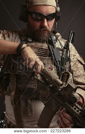 Brutal Man In The Military Desert Uniform And Body Armor Stands And Holds His Rifle On A Black Backg