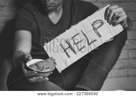 Homeless poor man holding carton board with word HELP and bowl for donations while sitting near brick wall