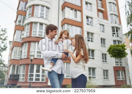 Low Angle Shot Of A Young Happy Family Posing In Front Of New Apartment Building. Father And Mother 