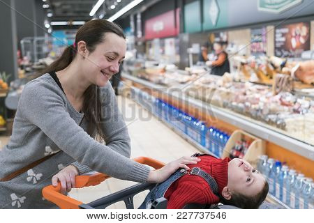 A Young Woman With A Small Son In A Supermarket. The Boy Sits In A Baby Cart And Laughs.