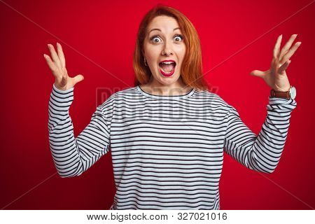 Young redhead woman wearing strapes navy shirt standing over red isolated background celebrating crazy and amazed for success with arms raised and open eyes screaming excited. Winner concept