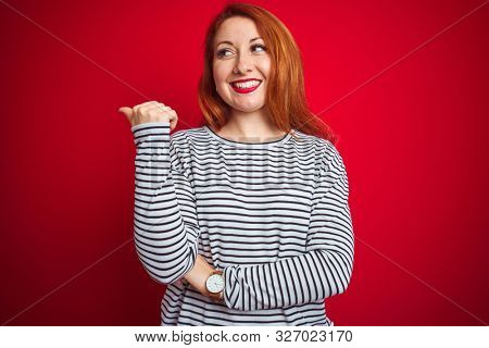 Young redhead woman wearing strapes navy shirt standing over red isolated background smiling with happy face looking and pointing to the side with thumb up.