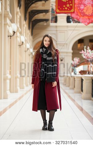 Full body portrait. Young beautiful woman in stylish in a long burgundy coat, indoor shop
