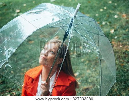 Caucasian Girl With Red Gabardine Walks Through A Park Under A Transparent Umbrella.