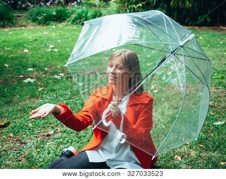 Caucasian Girl With Red Gabardine Walks Through A Park Under A Transparent Umbrella.