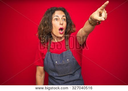 Middle age senior woman wearing apron uniform over red isolated background Pointing with finger surprised ahead, open mouth amazed expression, something on the front