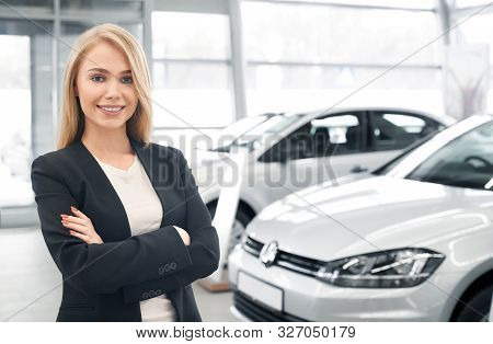 Smiling Female Car Dealer With Folded Arms Looking At Camera And Posing In Front Of New Automobiles.