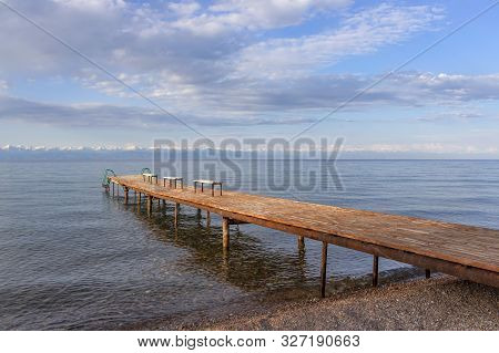 Pier With Benches On Lake Issyk-kul With A Mountain Range With Snow-capped Peaks On The Horizon Agai