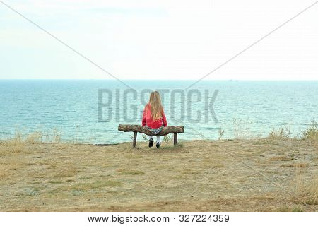 A Little Girl Sits Alone. Solo Travelling. A Tourist Sits On A Wooden Bench On A Cliff Above The Sea