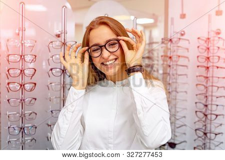 Happy Female Optometrist, Optician Is Standing With Many Glasses In Background In Optical Shop. Stan