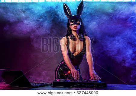 Young Sexy Woman Dj Playing Music In Mask. Headphones And Dj Mixer On Table. Smoke On Background