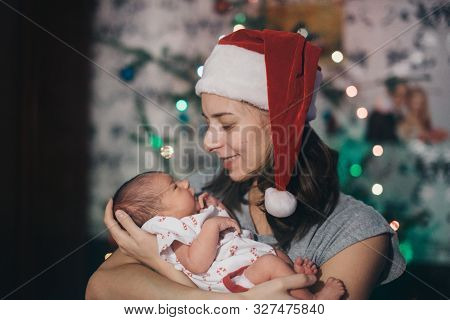 A Woman In A Christmas Hat Holding A Newborn Baby On The Background Of A Christmas Tree In Garlands.