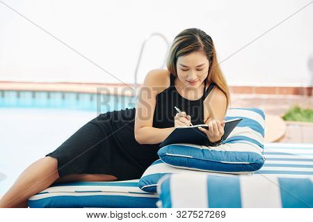 Beautiful Smiling Young Woman In Black Bodycon Dress Lying On Chaise-longe By The Pool And Filling G