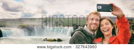 Selfie couple tourists taking photo with phone at nature famous Godafoss waterfall tourist destination, Iceland. Icelandic nature landscape. Interracial with cellphone couple having fun.