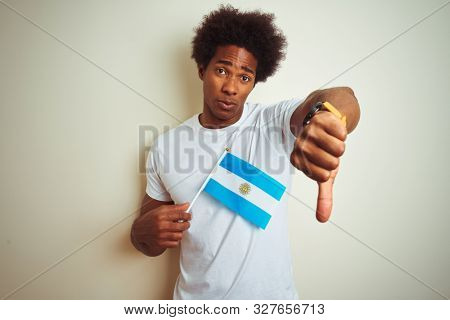 Afro american tourist man holding Argentinian Argentina flag over isolated white background with angry face, negative sign showing dislike with thumbs down, rejection concept