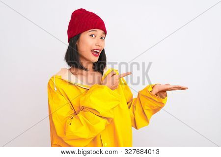 Young beautiful chinese woman wearing raincoat and wool cap over isolated white background amazed and smiling to the camera while presenting with hand and pointing with finger.