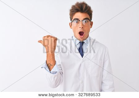 Young handsome sciencist man wearing glasses and coat over isolated white background Surprised pointing with hand finger to the side, open mouth amazed expression.