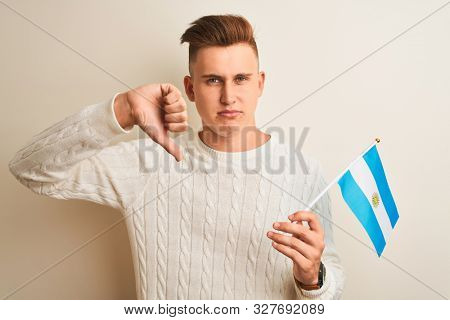 Young handsome man holding Argentinian Argentina flag over isolated white background with angry face, negative sign showing dislike with thumbs down, rejection concept