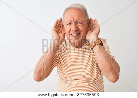 Senior grey-haired man wearing striped t-shirt standing over isolated white background Trying to hear both hands on ear gesture, curious for gossip. Hearing problem, deaf