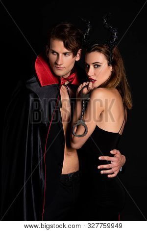 Handsome Man In Cloak Hugging Woman With Horns Holding Handcuffs Isolated On Black