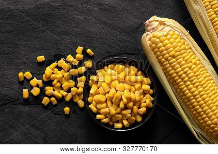 Canned Sweet Corn In A Black Ceramic Bowl Next To Two Corn Cobs In Husks And Spilled Corn On Black S