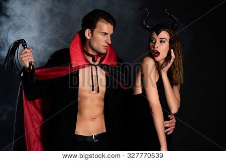 Handsome Man In Cloak Holding Flogging Whip Near Surprised Woman With Horns On Black With Smoke