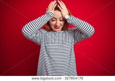 Young redhead woman wearing strapes navy shirt standing over red isolated background suffering from headache desperate and stressed because pain and migraine. Hands on head.