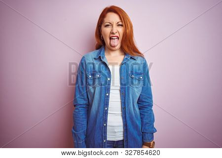 Youg beautiful redhead woman wearing denim shirt standing over isolated pink background sticking tongue out happy with funny expression. Emotion concept.