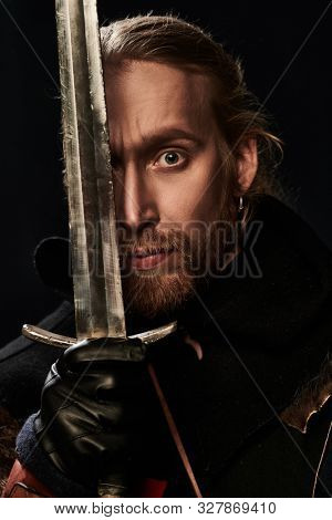 Close-up portrait of a medieval knight in armour and with a sword over black background. Historical reenactment.