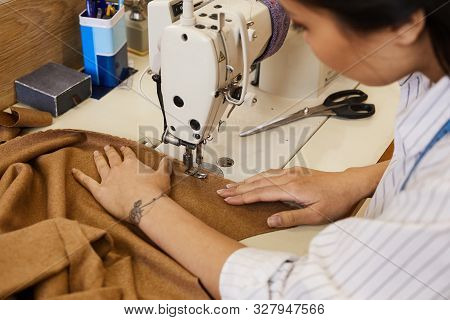 Close-up Of Young Woman Sewing Clothes From Brown Textile With The Help Of Sewing Machine In Worksop