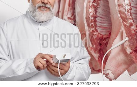 Close Up, Cropped Portrait Of Butcher With Gray Beard In White Uniform Near Pork Carcasses. Elderly 